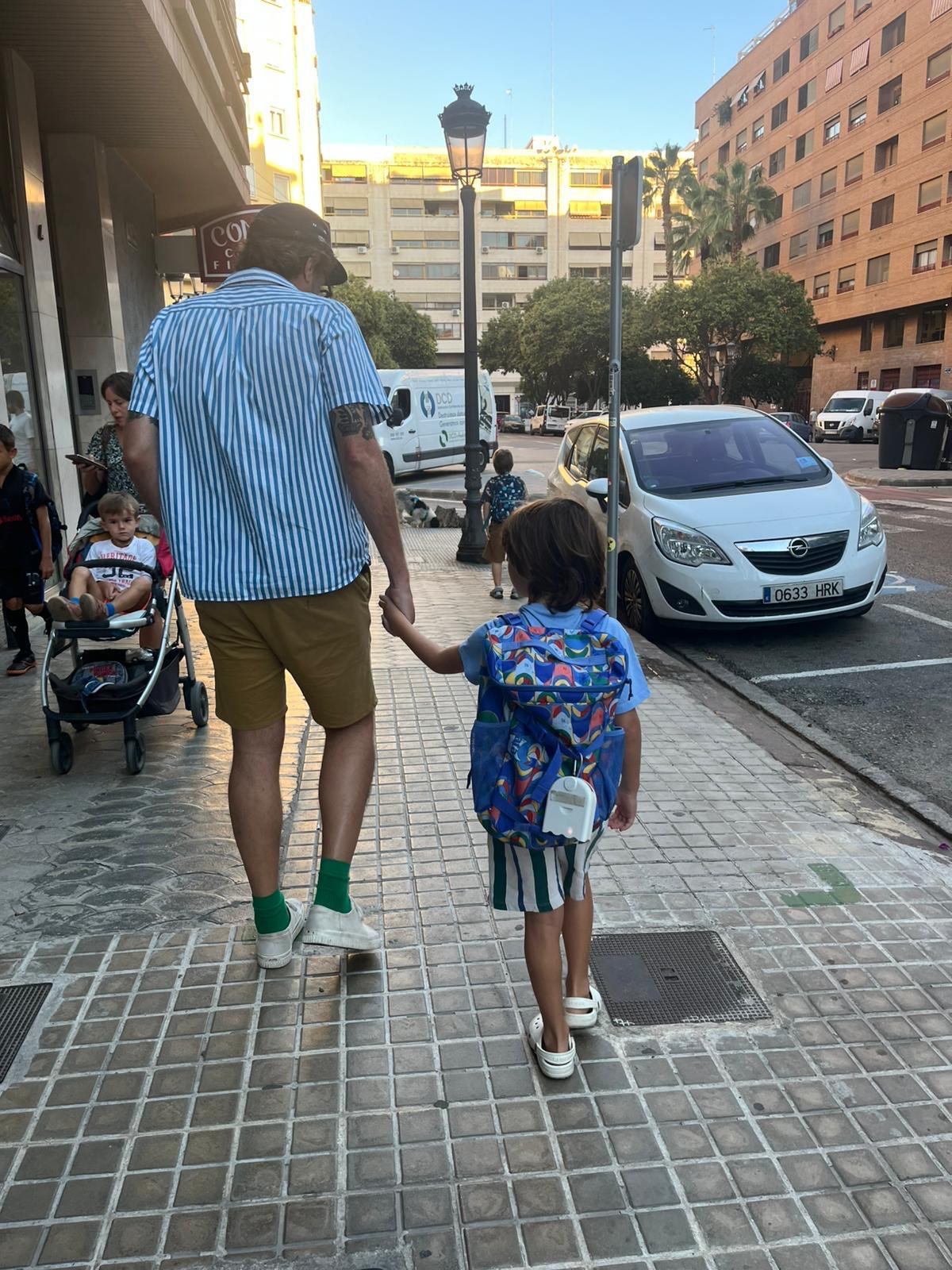 Fotografía voluntarias CEIP Cervantes Voluntarias del CEIP Cervantes andando de la mano por la acera de camino al colegio. El medidor de micropartículas se ve colgando de la mochila infantil escolar